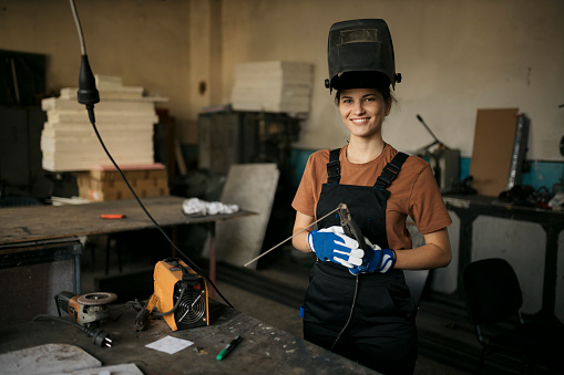 Portrait of a happy young woman skilled welder wearing protective apron, gloves and helmet standing in metal working workshop. Female welder working in a small metal shop looking at camera and smiling.
