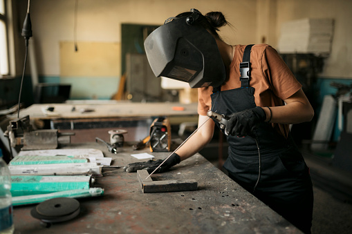 Female worker with safety helmet working with arc welding machine in metal workshop. Woman mechanical worker welding metal pieces on workbench at workshop.