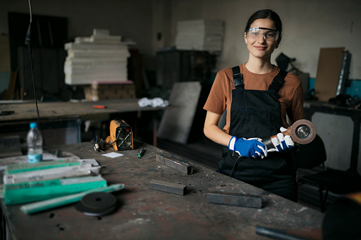 Portrait of young caucasian woman with safety equipment standing at workbench with a grinder in workshop. Young worker working in a small workshop looking at camera and smiling.
