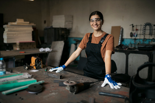 Portrait of a beautiful caucasian woman in protective workwear standing by workbench looking at camera with a smile. Confident young female wearing safety equipment looking at camera and smiling in small metal workshop.