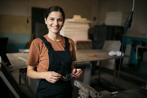 Portrait of a beautiful woman standing in metal working factory. Smiling caucasian woman holding safety glasses looking at camera standing in small metal workshop.