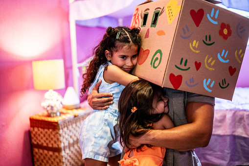 Father with a cardboard box on head embracing his daugther in the bedroom at home