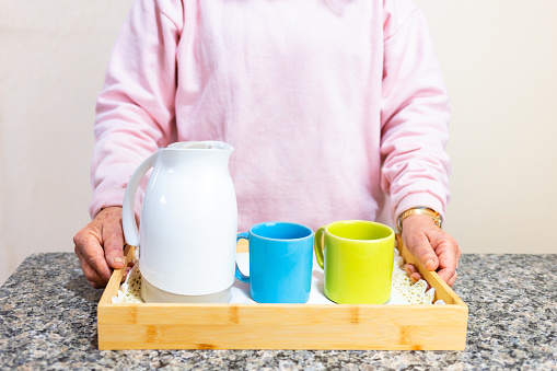 Two feminine hands holding a wooden tray, on which rests a coffee thermos and two porcelain mugs