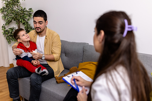 Handsome mid adult Caucasian single parent sitting at therapist with his son talking about problems