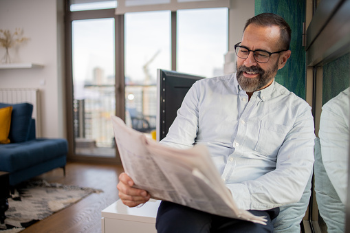 Businessman sitting on the couch and reading news on the financial newspaper, he is smiling at camera