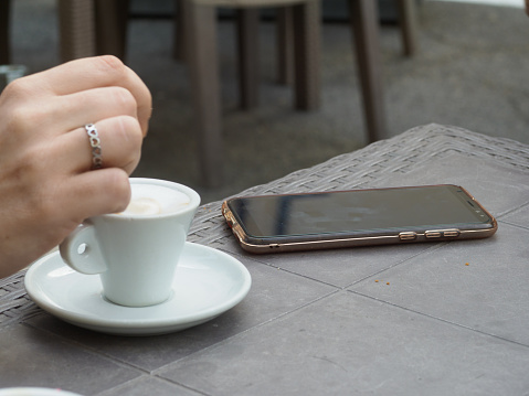 person having espresso coffee outdoors , stirring sugar with spoon, a smartphone on the table
