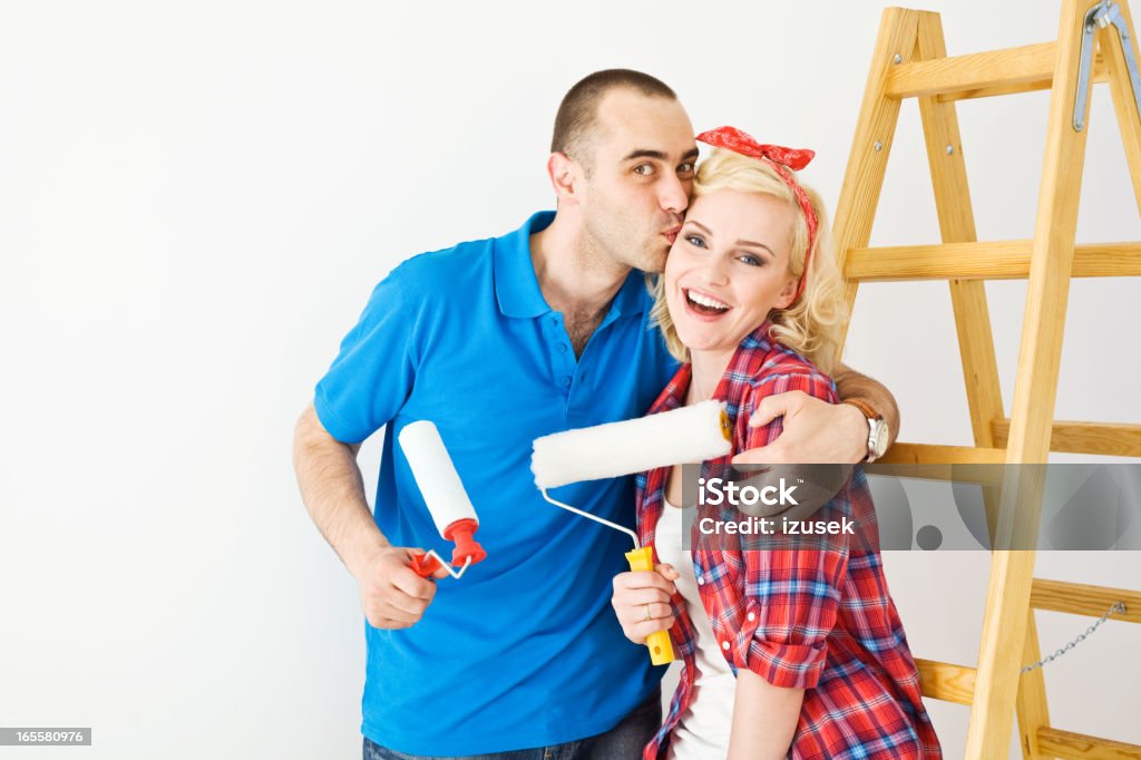 Happy couple painting a wall An attractive adult couple standing against white wall, next to the ladder and holding paint rollers in their hands. Looking at camera and smiling. A man kissing his wife. 25-29 Years Stock Photo