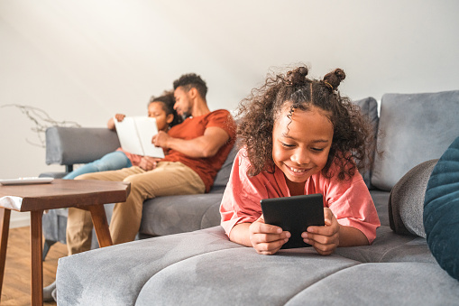 Mixed race girl lying down on a sofa in a domestic living room reading an e-book using e-reader. Sister and Black father reading a book in the background.