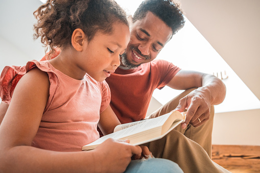 Black father and mixed mixed race girl sitting on a bed in a bedroom reading a book together.