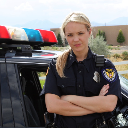 A caucasian female Police Officer with blond hair stands with her arms crossed in front of her police squad car.  Her uniform includes a badge, gun, and a two-way radio.
