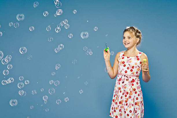 Cheerful, young 9 years old girl blowing soap bubbles with fun, studio shot, blue background, horizontal.