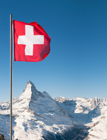 The white on red cross of the Swiss national flag flying above the European Alps, with the peak of the Matterhorn directly underneath the flag.  Taken at the ski resort of Zermatt.