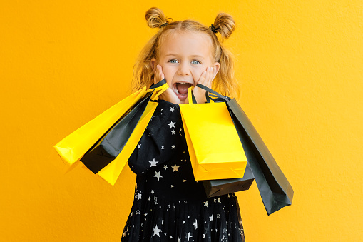 Surprise child girl open mouth hold black package bags purchases isolated yellow background studio. Shopping buy online discount sale. Cyber monday. Black friday. Toddler excited face kids emotions