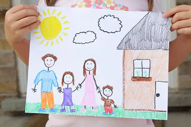 A four year old girl displays her drawing of her family standing outside of their beautiful new home.  The family includes a father, daughter, mother, and son all holding hands.  The drawing also includes a bright sun, clouds, and a house.