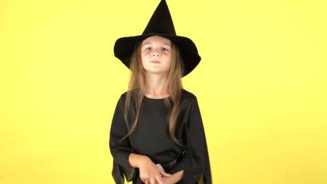 A little girl in a Halloween witch costume dances and shows emotions on a yellow studio background