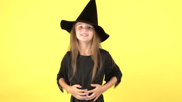 Happy Halloween concept. Portrait of little girl in witch costume raises hands scares and looking at camera, posing isolated on yellow color background wall in studio.