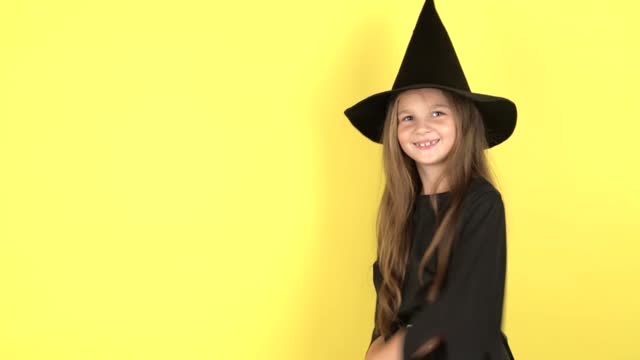 Little girl in witch costume , demonstrates copy space, points aside against yellow studio background for advertisement, dressed in Halloween costume.