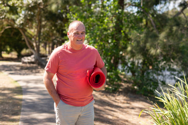 Overweight middle aged man walking to yoga Overweight middle aged man walking in a park with a yoga mat sports training australia stock pictures, royalty-free photos & images