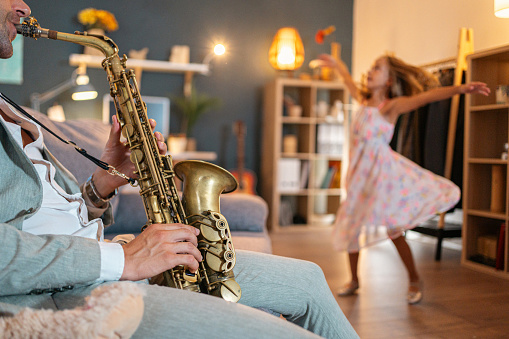 Adorable Caucasian girl dancing in the living room, while her father playing saxophone