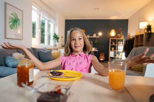 Adorable Caucasian girl, sitting for an dining table while eating jam or marmalade on a bread
