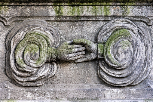 Two hands reaching out to each other from symbolic clouds on old stone carving covered in moss on 19th century cemetery