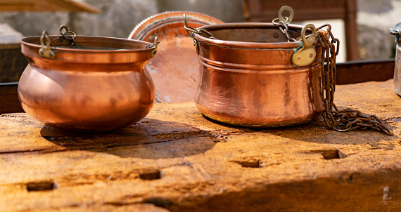 Close-up of antique shiny copper pots with rusty chains on wood-wormed table during vintage flea market in Italian small town