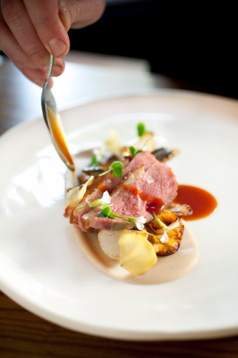 A chef at an upscale Bay Area restaurant using a spoon to drizzle sauce over slices of roasted duck breast.  The duck breast is beautifully arranged with chanterelles, turnips and edible flowers.