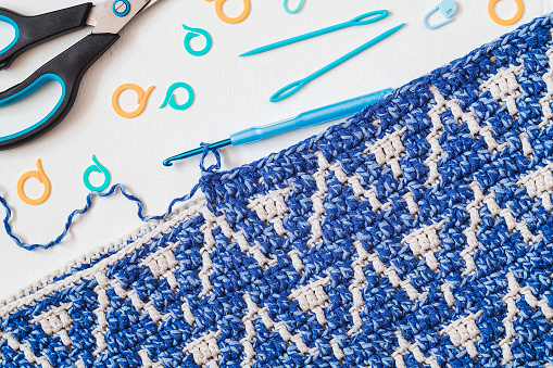 Blue white crochet fabric with crochet hook, scissors and knitting markers on a white background. Top view.