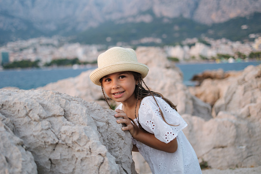 Cute little girl posing to photographer at sea coast. Beautiful little girl in white dress and knitted hat. High quality photo