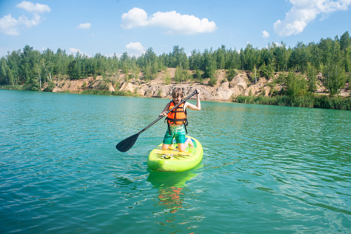 a boy rides a light green SUP board along a mountain river on summer vacation. A 10-year-old child goes in for water sports, in a life jacket, stands on a SUP surfboard.