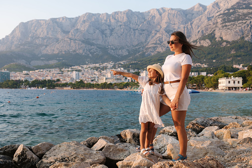 Beautiful mom and daughter stands on rocks and look at the sea. Back view of big mountains. High quality photo