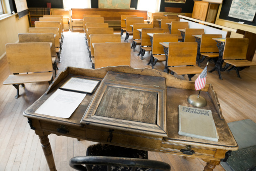 This is a horizontal, color photograph of an old fashioned school room out West. The point of view is from behind the teacher's desk. Rows of antique wooden school desks are placed in rows. 