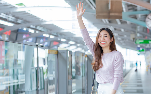 Asian beautiful lwoman smiling with happiness and refresh, wearing casual business shirt, waiving hand to greet, meet friend as appointment, standing on platform, waiting for sky train transportation in morning day