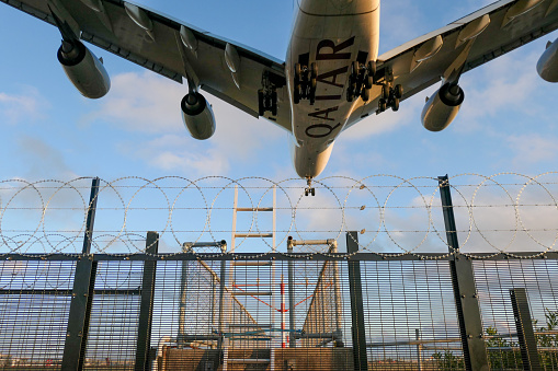 A Qatar Airways Airbus A380-861 plane, registration A7-APC, coming into land from the north on the main runway of Sydney Kingsford-Smith Airport as flight QR908 from Doha.  She is passing over a new security fence with razor wire, which protects one of the navigational landing light structures, visible through the wire and ladder.   This image was taken from near Nigel Love Bridge, Mascot on a sunny and partly cloudy afternoon at sunset on 2 September 2023.