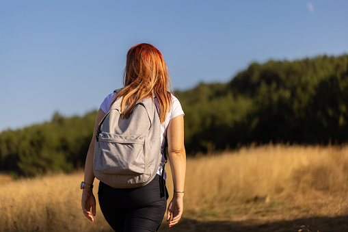 Rear view of unrecognizable mid-adult redhead Caucasian woman enjoying her hike through a wheatgrass field, during a majestic summer day in nature