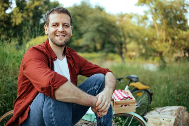 In the fall, a man sits on the grass, eats a sandwich with cheese and sausage and looks at a glass of coffee. A quick and tasty snack while walking in the fresh air. Selective focus. Close-up.