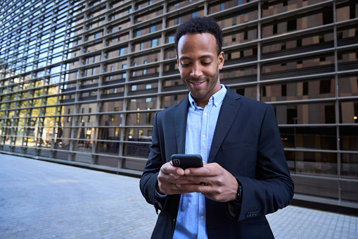Young adult Latin professional outside modern office building using phone. Business man in formal clothes typing message browsing online on mobile. Smiling entrepreneur consulting electronic agenda