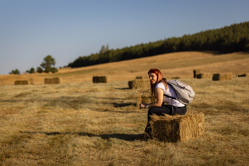 Carefree mid-adult redhead Caucasian woman sitting on a haystack, while enjoying the fresh and serene day in the nature