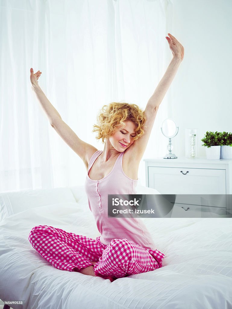 Happy morning Happy young adult woman sitting cross-legged on a bed in the morning and stretching her arms. Waking up Stock Photo