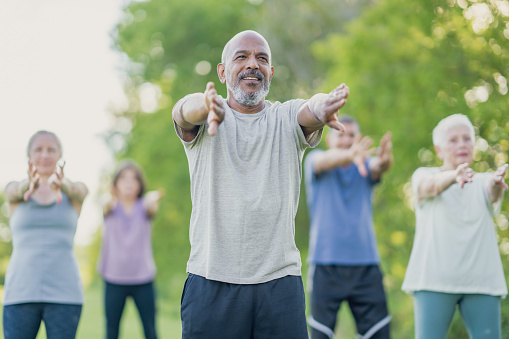 A small group of mature adults practice Tai Chi outdoors together on a warm summer day.  They are each dressed comfortably and are smiling as they take in the fresh air.