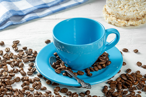 Morning coffee - an empty blue cup with a saucer for coffee and scattered coffee beans and rice cakes on a white wooden table.