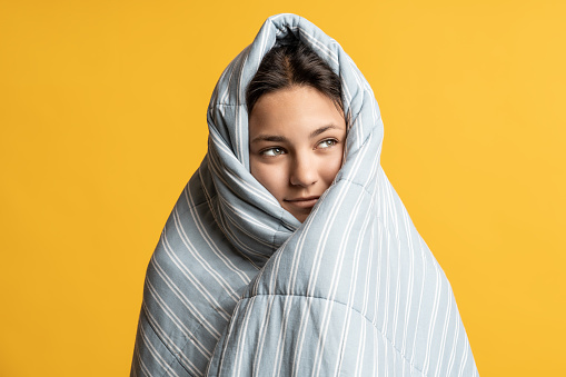 Teenager girl wrapped in warm blanket with head looking on copy space isolated on yellow background. Woman on advertisement banner, poster covering duvet. Bedtime, comfort, rest concept. Sleep product