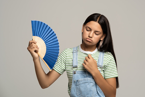Sweaty overheated teenage girl using paper fan suffer from heat feels sluggish. Displeased tired teen girl in overalls cooling in hot summer weather isolated on studio gray background. Stuffiness.
