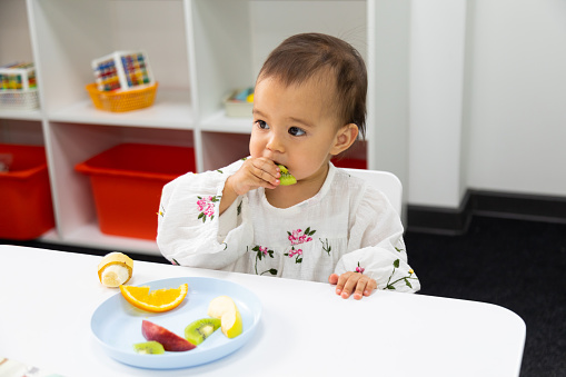 Closeup of toddler girl in beautiful dress eating cut fruits in the plate and sitting by the small table in kindergarten