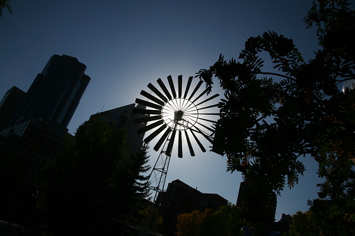 Silhouette of windmill with buildings and trees against a clear blue sky with the sun light in the center of windmill