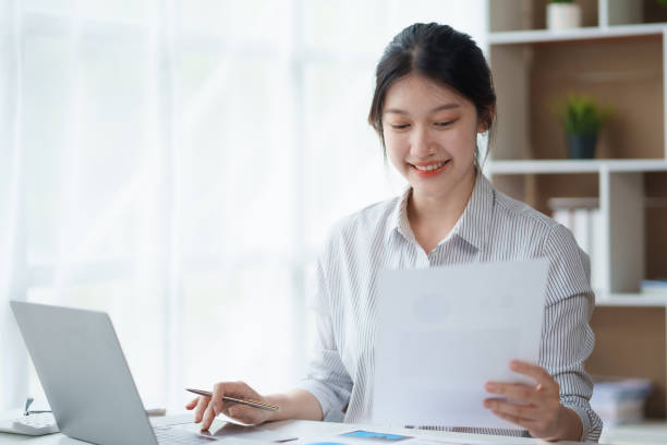istock Attractive asian businesswoman sitting at work using laptop and examining many documents in office smiling. 1657354071