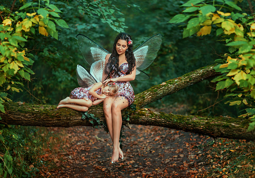 Fantasy woman fairy, sitting on log stroking with love sleeping little girl. Pixie is dreaming. Mom reads fairy tale to daughter, sings lullaby. family shooting motherhood care. trees summer forest.