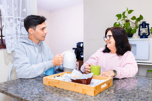 A mature woman and her adult son sit at a table, enjoying a traditional Brazilian afternoon coffee break. They engage in a lively and happy conversation amidst the backdrop of a typical Brazilian home setting