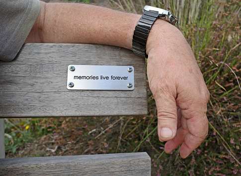 'Memories live forever' is written on a plaque on the back of a bench. A man's arm shelters it.
