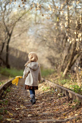 A little girl with a yellow maple leaf in her hand walks along the abandoned railway tracks. Back view. No face. Autumn lifestyle and outdoor recreation concept.